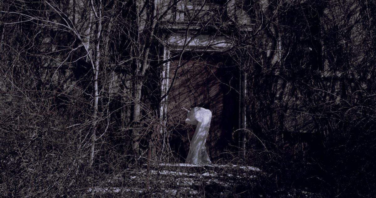 A pale, ghostly figure stands in the doorway of a ruined structure deep in bare winter woods — Disturbance